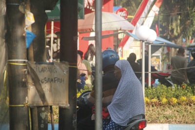 A person wearing a blue helmet is riding a motorcycle with a passenger who is wearing a polka-dotted headscarf. There are trees and a sign in the foreground, with a street scene in the background featuring people, plants, and a Coca-Cola sign.