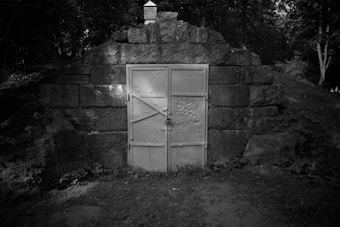 A heavy metal door is set into a stone wall in a wooded area. The door has numerous dents or marks, suggesting wear or possible bullet impacts. Surrounding foliage and trees indicate a forest setting, with shadows suggesting the presence of sunlight filtering through the leaves.