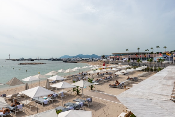 A sandy beach lined with numerous white lounge chairs and sun umbrellas. Few people are relaxing under the umbrellas. There are palm trees and various colorful flags along a beachfront promenade. The calm sea stretches into the distance with several yachts docked at a marina. Mountains are visible in the background under a slightly overcast sky.