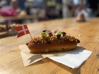 A hot dog topped with crispy fried onions and diced vegetables is placed on a paper napkin on a wooden table. The hot dog is garnished with a small Danish flag. The setting appears to be outdoors with blurred people in the background suggesting a casual, social atmosphere.