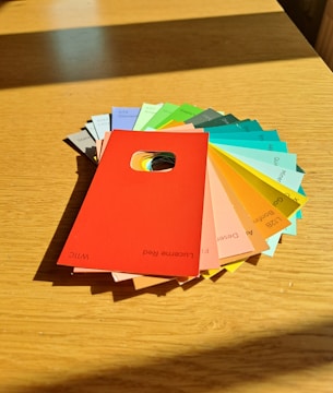 A collection of colorful paint swatches arranged on a wooden table. The swatches fan out, showing a variety of colors including red, orange, yellow, green, blue, and grey. Each swatch has a written name and code at the bottom, and the central swatch is a vivid red color with a rectangular hole near the center.