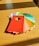 A collection of colorful paint swatches arranged on a wooden table. The swatches fan out, showing a variety of colors including red, orange, yellow, green, blue, and grey. Each swatch has a written name and code at the bottom, and the central swatch is a vivid red color with a rectangular hole near the center.