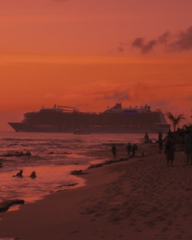 A large cruise ship moves across the water at sunset, casting a silhouette against the vibrant orange and pink sky. The foreground shows a sandy beach with several people walking along the shore and a couple swimming in the ocean. Palm trees are visible, adding to the tropical atmosphere.