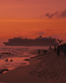 A large cruise ship moves across the water at sunset, casting a silhouette against the vibrant orange and pink sky. The foreground shows a sandy beach with several people walking along the shore and a couple swimming in the ocean. Palm trees are visible, adding to the tropical atmosphere.