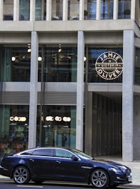 A modern building with large glass windows and a concrete facade. A black luxury sedan is parked in front of the building. The illuminated circular sign in one window reads 'Jamie in Victoria Oliver.' There are lights on inside the building and visible reflections on the glass.