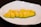 A neatly arranged row of diced mango segments is placed on a clean, white plate. The mango pieces are vibrant yellow with hints of green on the edges, suggesting freshness and ripeness. The background is a dark surface, providing contrast to the plate.