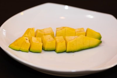 A neatly arranged row of diced mango segments is placed on a clean, white plate. The mango pieces are vibrant yellow with hints of green on the edges, suggesting freshness and ripeness. The background is a dark surface, providing contrast to the plate.