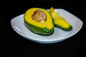 A halved avocado with its seed is placed on a white plate. The interior is a vivid yellow-green, contrasting with the darker green of the skin. One piece is sliced, showing the smooth texture of the fruit.