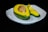 A halved avocado with its seed is placed on a white plate. The interior is a vivid yellow-green, contrasting with the darker green of the skin. One piece is sliced, showing the smooth texture of the fruit.