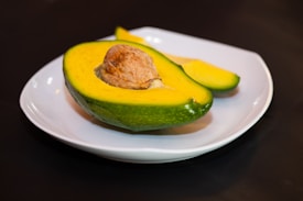 A halved avocado with its brown seed resting on a white plate. The avocado is ripe, displaying a vibrant green skin and yellowish-green flesh. A slice of avocado is positioned next to the halved piece, all set against a dark, contrasting background.