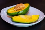 A ripe, halved avocado with its seed visible, placed on a white plate. The flesh of the avocado is bright yellow with hints of light green, and the outer skin is a rich green. The avocado is set against a dark background, highlighting its vibrant colors.
