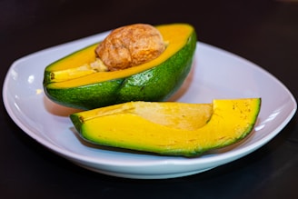A ripe, halved avocado with its seed visible, placed on a white plate. The flesh of the avocado is bright yellow with hints of light green, and the outer skin is a rich green. The avocado is set against a dark background, highlighting its vibrant colors.