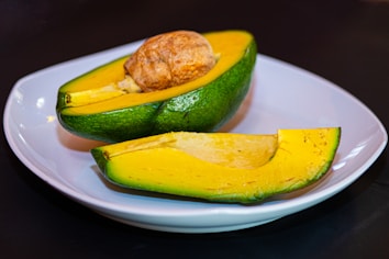A ripe, halved avocado with its seed visible, placed on a white plate. The flesh of the avocado is bright yellow with hints of light green, and the outer skin is a rich green. The avocado is set against a dark background, highlighting its vibrant colors.