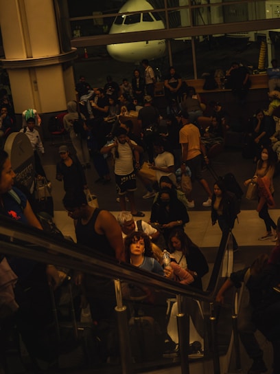 A bustling airport terminal with a diverse crowd of travelers, some seated and others standing. An airplane is visible through the large windows in the background. People appear to be waiting with luggage, some using their phones, while others are engaged in conversation. The lighting is dim, giving the scene a subdued atmosphere.