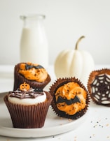 Three Halloween-themed cupcakes are displayed on a white plate. Each cupcake is decorated with orange and black toppings, featuring designs such as bats, spider webs, and a miniature pumpkin topper. A small white pumpkin and a bottle of milk in the background add to the festive setup.