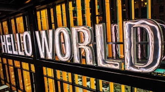 Large neon letters spell out 'Hello World' against a backdrop of vertical orange and black strips. The letters are illuminated, creating a bright and inviting ambiance.