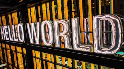 Large neon letters spell out 'Hello World' against a backdrop of vertical orange and black strips. The letters are illuminated, creating a bright and inviting ambiance.