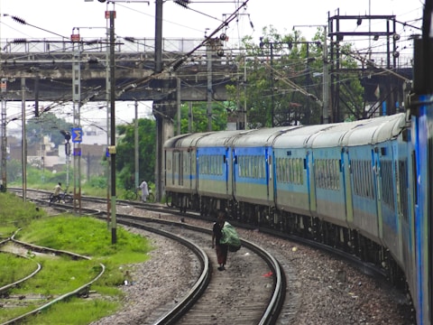 A train with blue and gray carriages is traveling along curved railway tracks under an overpass with electrical lines. The surroundings include greenery and a few buildings in the background. People are seen walking or standing near the tracks, and bicycles are present.