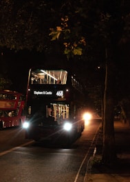 A double-decker bus is illuminated by its headlights as it travels on a dimly lit street at night. The destination sign reads 'Elephant & Castle 468'. Another similar bus is visible in the background on the left side of the street. The scene is surrounded by shadows from trees and streetlights.