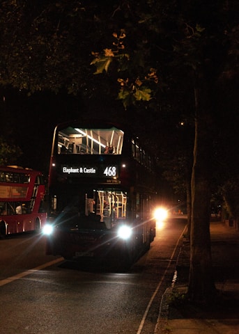 A double-decker bus is illuminated by its headlights as it travels on a dimly lit street at night. The destination sign reads 'Elephant & Castle 468'. Another similar bus is visible in the background on the left side of the street. The scene is surrounded by shadows from trees and streetlights.