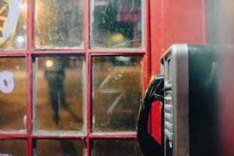 A vintage telephone booth with a red frame and a black telephone inside. The glass panels show reflections and graffiti. Dim lighting creates a nostalgic atmosphere.