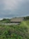 A rustic, thatched-roof cottage sits nestled amidst lush green vegetation. The structure is partially obscured by surrounding tall grass and foliage, with a wooden fence in front. The sky above is overcast, creating a moody backdrop to the serene landscape.