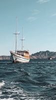 A large wooden sailing ship with two masts is navigating through a vast body of water. The waves are gentle, and the sky is clear with few clouds. In the background, there are rugged, mountainous islands.