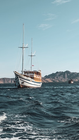 A large wooden sailing ship with two masts is navigating through a vast body of water. The waves are gentle, and the sky is clear with few clouds. In the background, there are rugged, mountainous islands.