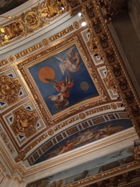 A richly decorated ceiling with ornate gold designs and intricate patterns. The central painting features angelic figures, one with red robes and another with white wings, set against a deep blue background with a large orange circle. The surrounding area is adorned with gold embellishments and classical motifs.