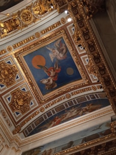 A richly decorated ceiling with ornate gold designs and intricate patterns. The central painting features angelic figures, one with red robes and another with white wings, set against a deep blue background with a large orange circle. The surrounding area is adorned with gold embellishments and classical motifs.