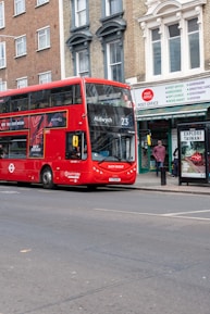 A red double-decker bus with the number 23 and destination Aldwych displayed on the front is parked on the side of a street. The street features a post office with a sign above its entrance. There are buildings with brick facades and tall windows. A bus stop advertisement promotes tourism, and a person stands nearby.