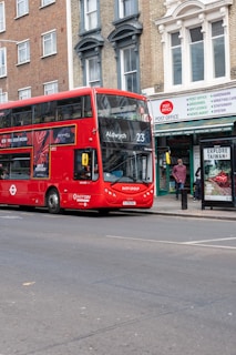 A red double-decker bus with the number 23 and destination Aldwych displayed on the front is parked on the side of a street. The street features a post office with a sign above its entrance. There are buildings with brick facades and tall windows. A bus stop advertisement promotes tourism, and a person stands nearby.