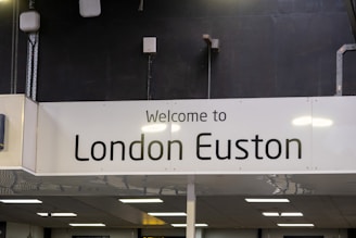 A sign with the text 'Welcome to London Euston' displayed against a dark background. The sign is rectangular and white, with modern lighting and various electrical installations visible in the surrounding area.