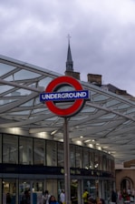 A London Underground sign prominently displayed against a modern glass and metal structure. The sky is overcast, and various people are seen walking around. A historic building with a spire is visible in the background.