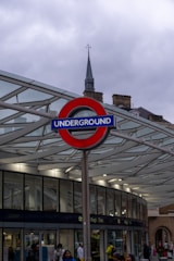 A London Underground sign prominently displayed against a modern glass and metal structure. The sky is overcast, and various people are seen walking around. A historic building with a spire is visible in the background.