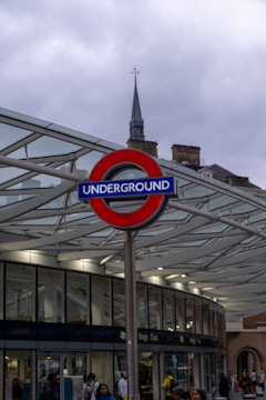 A London Underground sign prominently displayed against a modern glass and metal structure. The sky is overcast, and various people are seen walking around. A historic building with a spire is visible in the background.