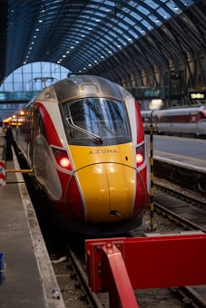 A modern passenger train with a sleek red and yellow design is stationed on train tracks inside a large, covered platform area. The station features an arched glass and steel canopy, and the platform is equipped with various signs and barriers. The train's headlights are on, and there are some lights illuminating the station.
