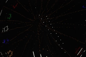 A dark night sky is dotted with multicolored stars and illuminated light structures forming patterns. The design creates a sense of depth and movement with its star-like formations spreading outward.