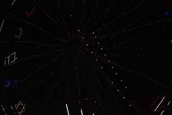 A dark night sky is dotted with multicolored stars and illuminated light structures forming patterns. The design creates a sense of depth and movement with its star-like formations spreading outward.