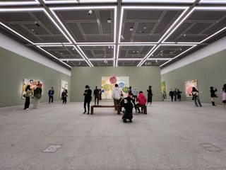A spacious art gallery with high ceilings and minimalistic design. People are observing colorful abstract paintings displayed on the walls. The lighting is bright and evenly distributed, highlighting the artwork.