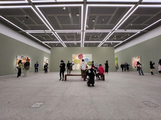 A spacious art gallery with high ceilings and minimalistic design. People are observing colorful abstract paintings displayed on the walls. The lighting is bright and evenly distributed, highlighting the artwork.