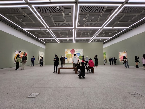 A spacious art gallery with high ceilings and minimalistic design. People are observing colorful abstract paintings displayed on the walls. The lighting is bright and evenly distributed, highlighting the artwork.