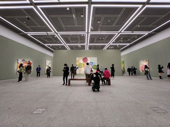 A spacious art gallery with high ceilings and minimalistic design. People are observing colorful abstract paintings displayed on the walls. The lighting is bright and evenly distributed, highlighting the artwork.