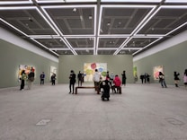 A spacious art gallery with high ceilings and minimalistic design. People are observing colorful abstract paintings displayed on the walls. The lighting is bright and evenly distributed, highlighting the artwork.