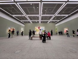 A spacious art gallery with high ceilings and minimalistic design. People are observing colorful abstract paintings displayed on the walls. The lighting is bright and evenly distributed, highlighting the artwork.
