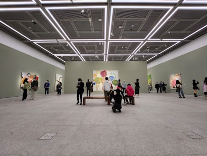 A spacious art gallery with high ceilings and minimalistic design. People are observing colorful abstract paintings displayed on the walls. The lighting is bright and evenly distributed, highlighting the artwork.