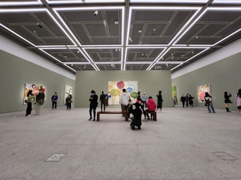 A spacious art gallery with high ceilings and minimalistic design. People are observing colorful abstract paintings displayed on the walls. The lighting is bright and evenly distributed, highlighting the artwork.