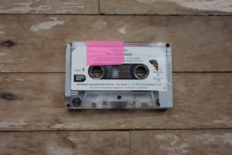 A transparent cassette tape resting on a wooden surface. The tape features a pink label with writing and contains printed text including 'Der Alchemist.' The casing reveals two spools visible through the transparent plastic, and the wood has a natural, weathered appearance with visible grain patterns.