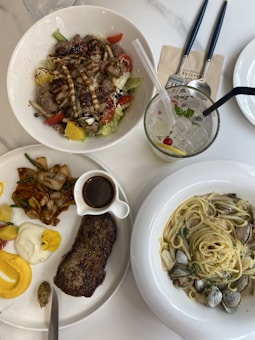 A meal setup featuring three different dishes placed on a white table. The top left bowl contains a colorful salad with grilled meat strips, cherry tomatoes, lettuce, and other vegetables. The bottom left plate includes a grilled steak with sides of roasted vegetables, sauces, and condiments. The right plate holds a serving of spaghetti with clams and herbs. Additionally, there is a glass of lemonade with ice, mint, and a straw, along with utensils wrapped in a napkin.