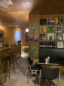 A cozy, dimly-lit bar with wooden interiors features a collection of vinyl records displayed on the wall. The seating area includes black tables and chairs. Posters and a variety of spirits and wine bottles are arranged on shelves. Warm lighting creates an inviting atmosphere.